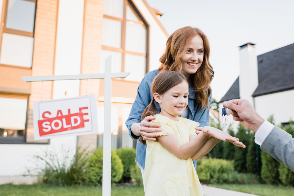Happy mother hugging daughter taking keys from broker near sign with sold lettering on blurred background
