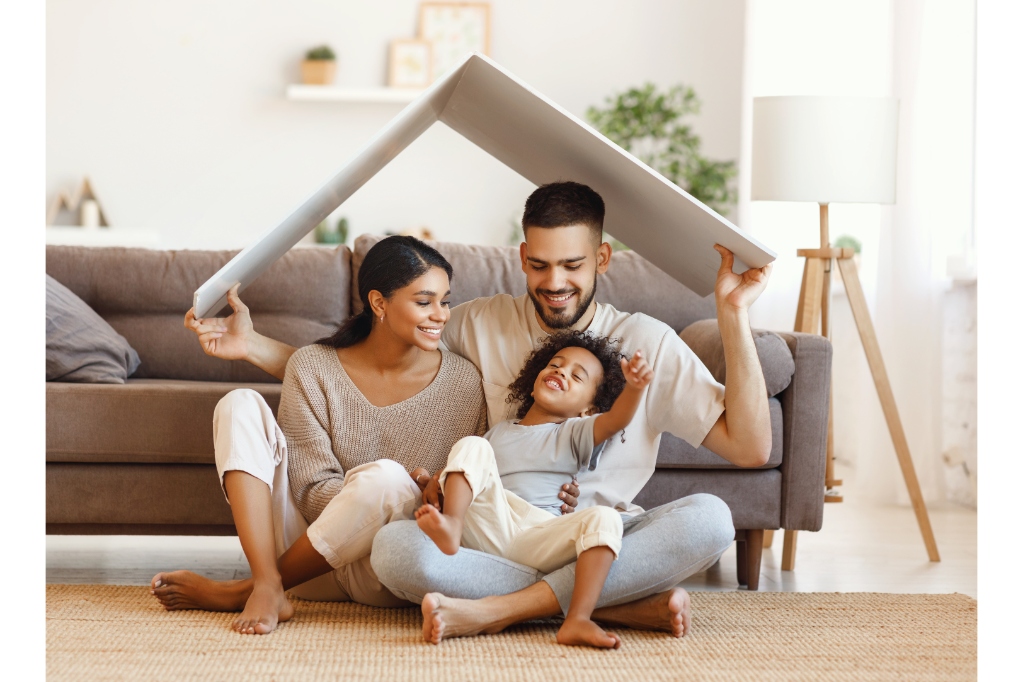 Delighted diverse parents and mixed race son sitting under roof and smiling near sofa in cozy living room