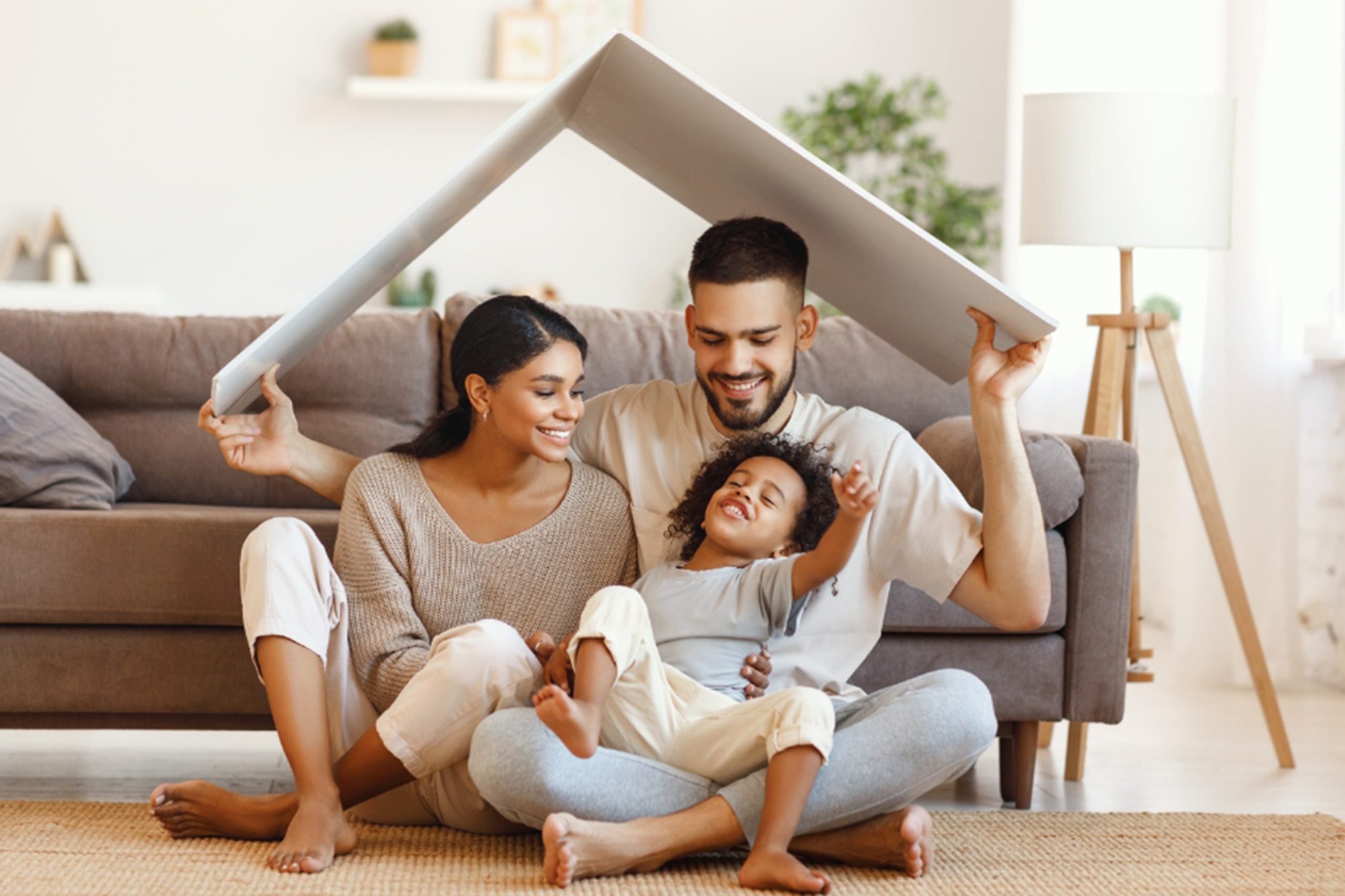 Delighted diverse parents and mixed race son sitting under roof and smiling near sofa in cozy living room